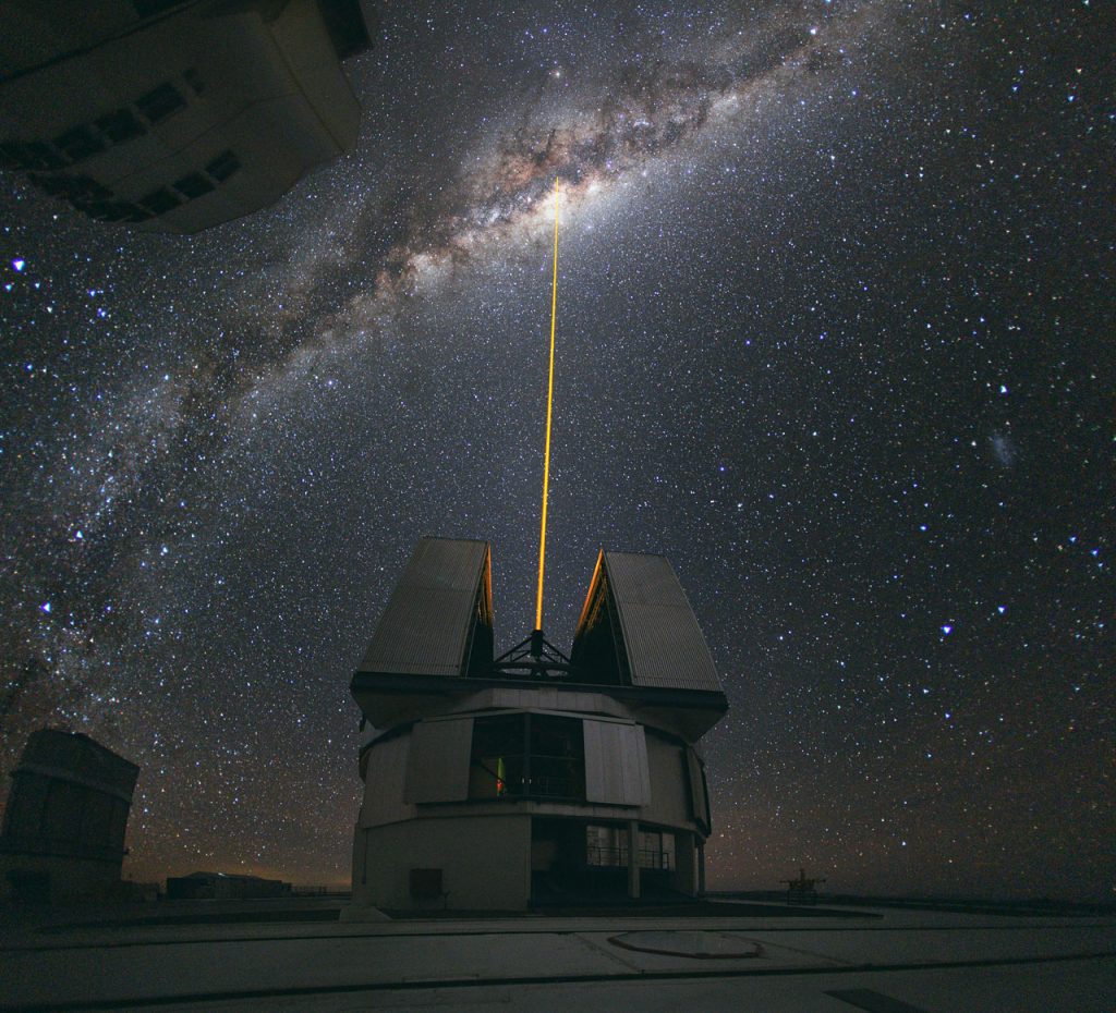 Photo montrant un des quatre télescopes principaux du VLT, le Yepun. Un laser jaune en part et pointe vers le ciel. Il sert de référence pour corriger les perturbations de l’atmosphère par optique adaptative.