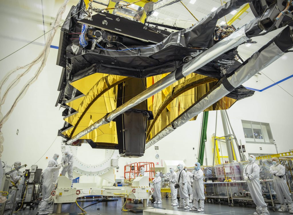 Le JWST en salle blanche en Californie. Il est suspendu en l'air au-dessus des ingénieurs en blouse blanche qui participent à son test et à son intégration.