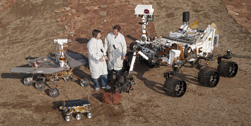 Photo couleur des maquettes en taille réelle des trois rovers Sojourner (premier plan), Opportunity (à gauche) et Curiosity (à droite) aux côtés de deux ingénieurs du Jet Propulsion Laboratory de la Nasa (Californie) où ils ont été développés. 