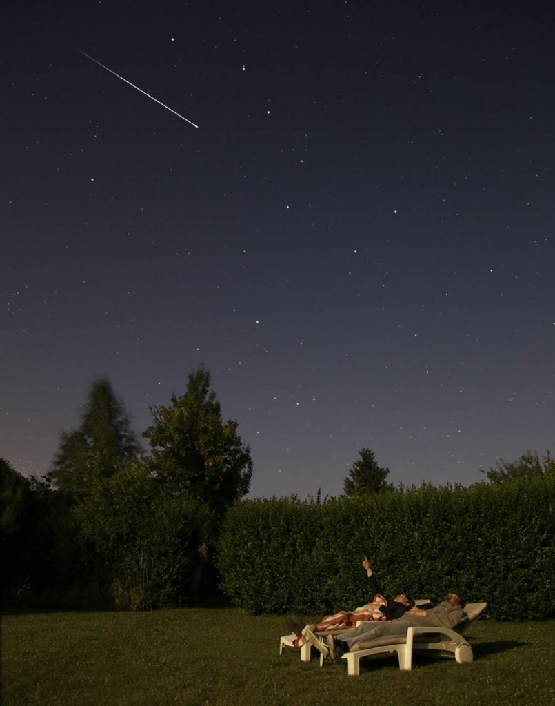 Photo montrant deux observateurs d'étoiles filantes sur des chaises longues. Ils sont dans un jardin et au dessus d'aux, sur un fond de ciel étoilé, une étoile filante de couleur blanche traverse le ciel de gauche à droite. L'un des observateurs montre l'étoile filante du doigt.