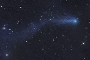 Comète PanSTARRS qui se déplace vers la droite de l'image, laissant derrière elle une traînée brillante très évasée, sur un fond de ciel étoilé.