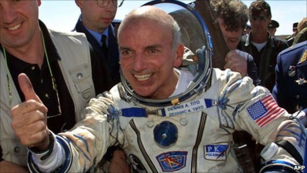 Dennis Tito, le premier touriste spatial à son retour en 2001, habillé de sa combinaison spatiale, un grand sourire et pouce levé.