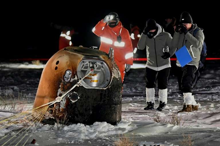 Photo de la sonde Chang'e 5 après son retour sur Terre. On la voit dans la neige, la nuit, capsule brûlée de couleur marron, entourée de trois scientifiques et ingénieurs habillés chaudement.  