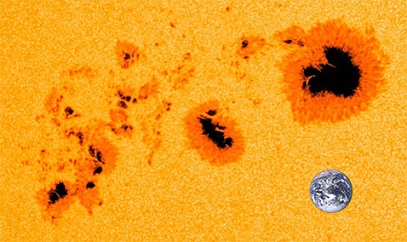 Zoom sur une partie du Soleil jaune clair où l'on voit trois principales tâches solaires (cœur noir et contour orange). Une petite Terre permet de se rendre compte de l'échelle : les tâches sont du même ordre de grandeur que notre planète.