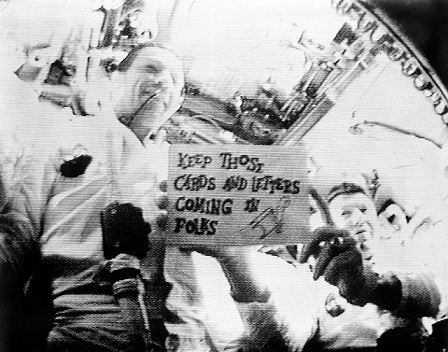Capture d'écran en noir et blanc de la première diffusion télévisée depuis l'espace dans laquelle les astronautes d'Apollo 7 brandissent un panneau indiquant "Continuez à nous envoyer des cartes et des messages".