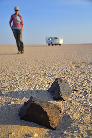 Illustration de la chasse aux météorites : deux météorites sont posées au premier plan sur le sol jaune du désert du Dhofar (Oman), derrière en flou une femme s'approche, elle est descendue d'un 4x4 aux portes ouvertes au troisième plan, aussi flou.