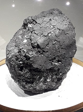 Photo de la météorite d'Orgueil, sur un support blanc, elle est ovoïdale et gris anthracite. 