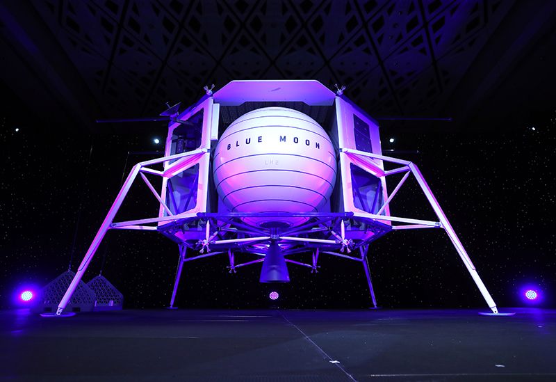 Sur une estrade, de couleur blanche et éclairé par des lumières bleu-violet, le concept d'atterrisseur lunaire de Blue Origin avec une sphère en son centre.