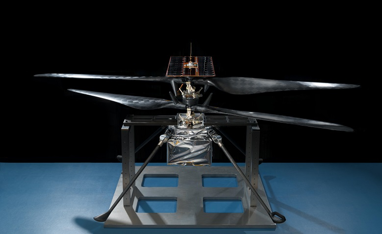 Photo du modèle de vol du Mars Helicopter Scout, l'hélicoptère qui va embarquer sur Mars 2020. Il est posé sur un socle bleu, au centre de l'image, on voit bien ses 4 pales. 