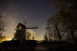 Photo d'un ciel de nuit avec la constellation d'Orion bien visible. Au sol en bas à gauche un moulin, en bas à droite de grands arbres.