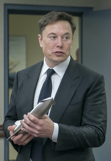 Photo portrait d'Elon Musk, patron de SpaceX, en costume gris foncé avec cravate, une maquette de navette spatiale à la main.