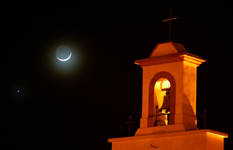 La planète Vénus se trouve à côté de la Lune en fin croissant, et à droite on voit un clocher occitan éclairé par des lampadaires