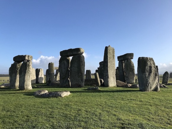 Photo du mégalithe de Stonhenge en Angleterre. On y voit plusieurs gros blocs de pierre gris, plantés dans le sol, au soleil, avec de l'herbe verte sur la partie basse de l'image et du ciel bleu sur la partie haute, les pierres au milieu.