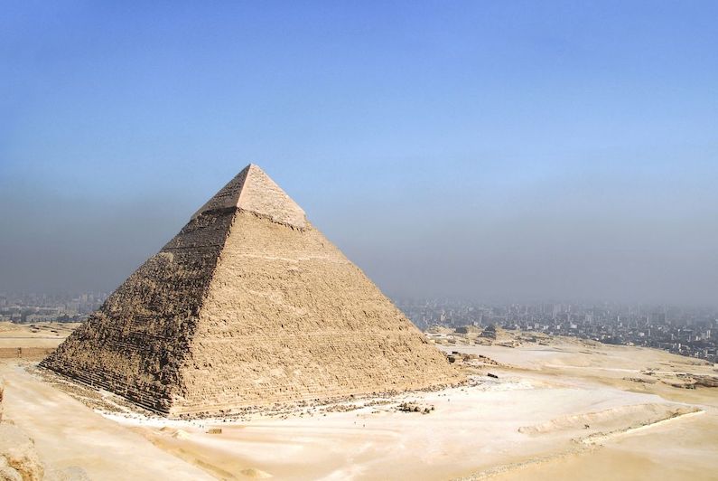 Pyramide de Khéops au soleil, avec la ville du Caire en fond sous un ciel bleu taché d'une nuage de pollution. 