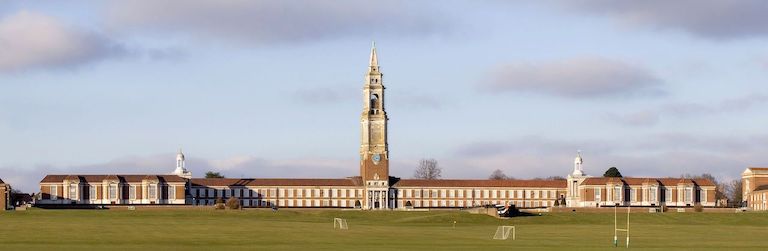 Photo de la Royal Hospital School à Greenwich : on voit au premier plan de la pelouse avec terrains de football et rugby, au centre le bâtiment en longueur en pierres beige foncé, fenêtres et volets blancs, avec un clocher au centre. Et en fond le ciel bleu avec des nuages rose et violet. 