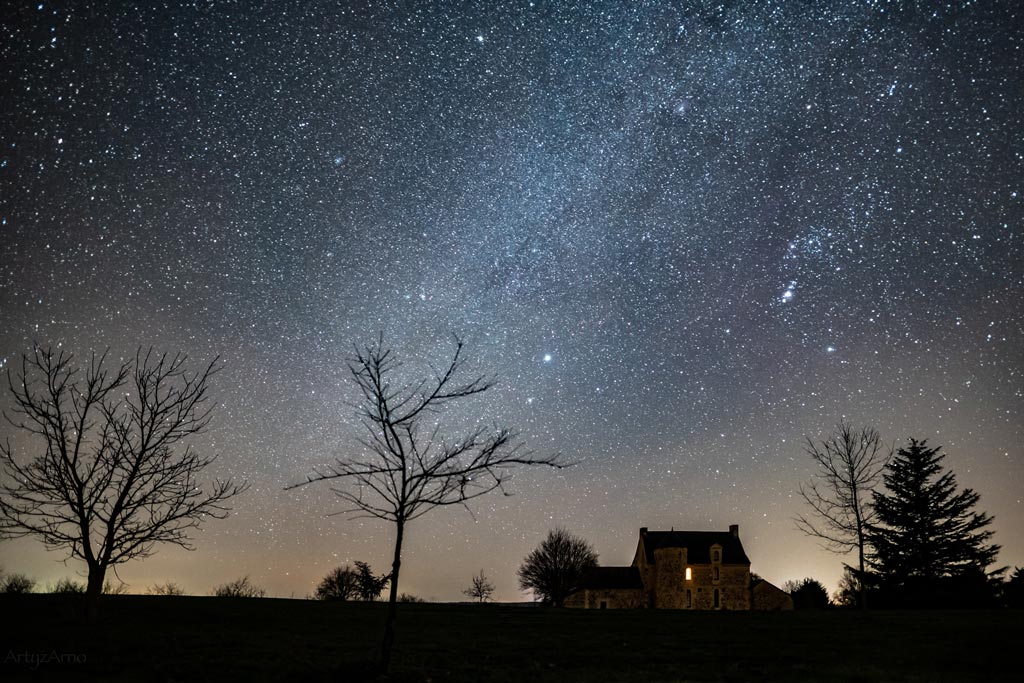 Paysage nocturne avec le ciel étoilé en hiver et la constellation d'Orion et en avant-plan des arbres et une maison.