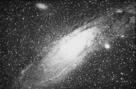 Première photo de la galaxie d'Andromède en noir et blanc. On voit la galaxie spirale inclinée d'en bas à gauche de l'image à en haut à droite, le centre complètement blanc, sur fond de ciel noir étoilé.