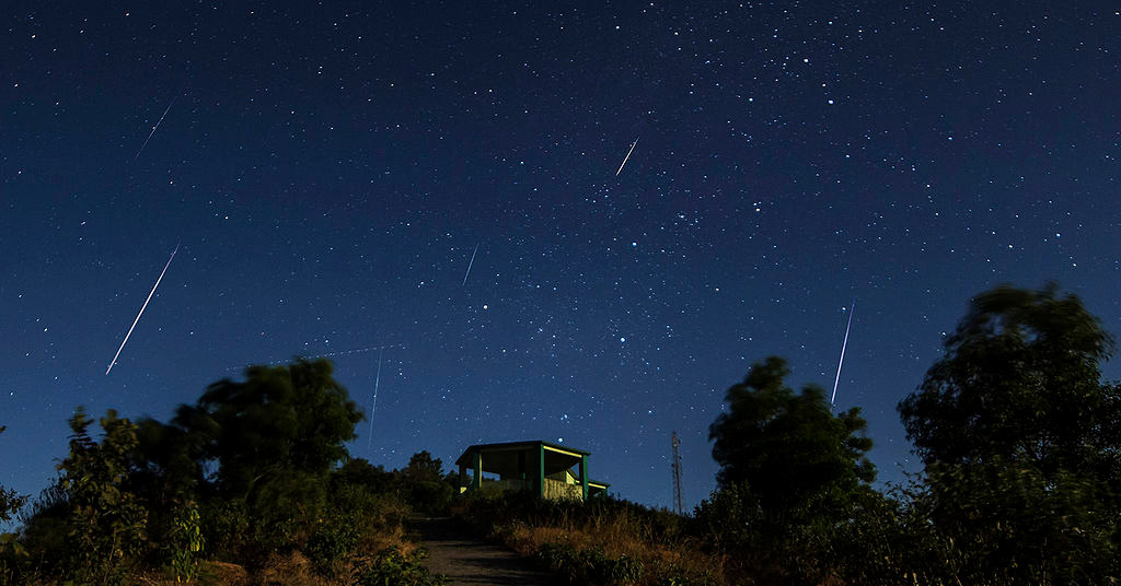 Ciel nocturne avec plusieurs traits blancs qui sont la trace du passage d'étoiles filantes. En premier plan, une colline et une cabane.