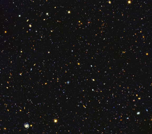 Sur fond noir, une multitudes de points et formes brillants, légèrement bleu, jaune, rouge, orange, vert... Ce sont 15 000 galaxies visibles sur le même cliché. En bas à gauche, on voit nettement une galaxie spirale. 