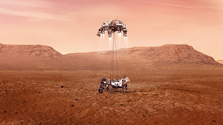 Le sol martien en couleur rouge orangé avec au loin des montagnes. Au milieu de l'image le rover Perseverance qui vient de toucher le sol, il est encore suspendu à l'étage de descente (capsule avec rétrofusées) par des filins.