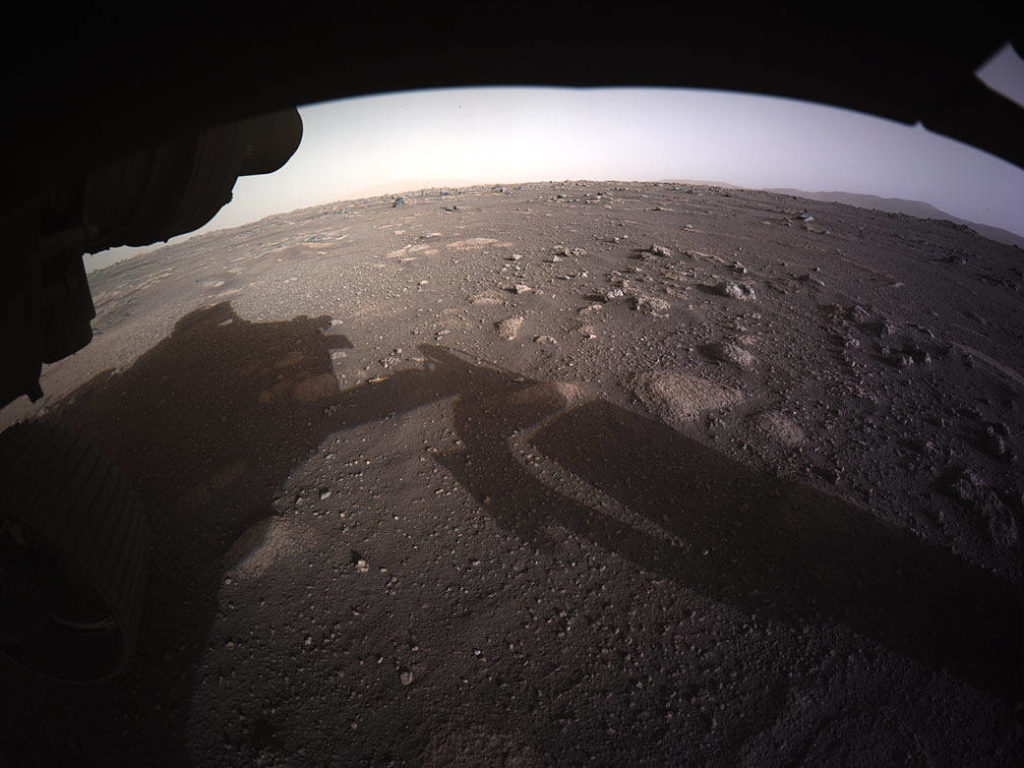 Première image du rover où l'on voit son ombre sur le sol, l'horizon martien sous une forme arrondie par l'effet de caméra. Le sol martien est en couleur marron et parsemé de petites bosses et de divers cailloux.  