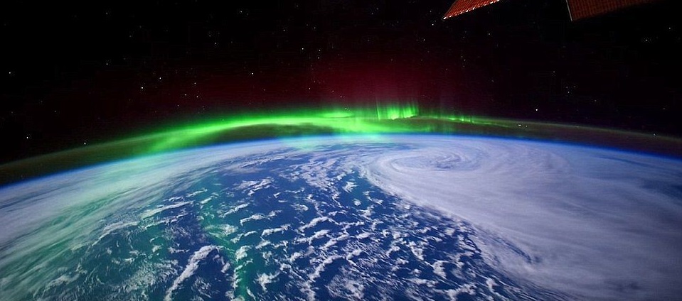 Image satellite d'une aurore boréale au-dessus du Canada : on voit la courbure de la Terre, le bleu de l'océan et des nuages blancs, et au-dessus, des rayons verts qui suivent cette courbure, avec en fond un ciel noir étoilé.