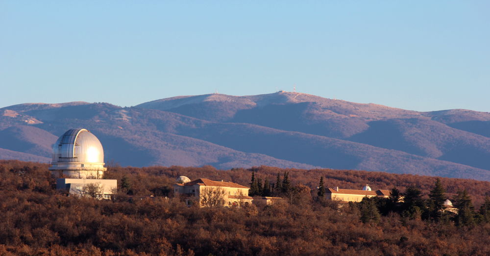 En début de journée, vue sur l'Observatoire de Haute-Provence avec en fond de la moyenne montagne et une végétation de mi-saison, un ciel bleu et au premier plan deux bâtiments de style provençal ainsi qu'à gauche une coupole argentée. Le Soleil éclaire la scène par la droite. 