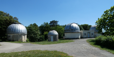 Sous un ciel bleu, trois coupoles de l'Observatoire de Lyon ensoleillées (toits bleu clair et murs blancs), séparées par un chemin goudronné et des surfaces d'herbe verte. 