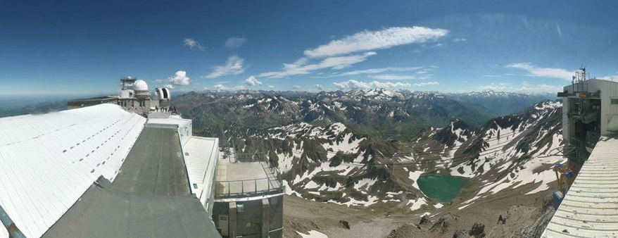 Vue panoramique depuis le Pic du Midi. De gauche à droite on voit le toit d'un des bâtiments, les coupoles blanches, au centre les montagnes avec un peu de neige et un lac bleu vert en contre-bas, le ciel est bleu, et à droite de l'image une autre partie des bâtiments. 