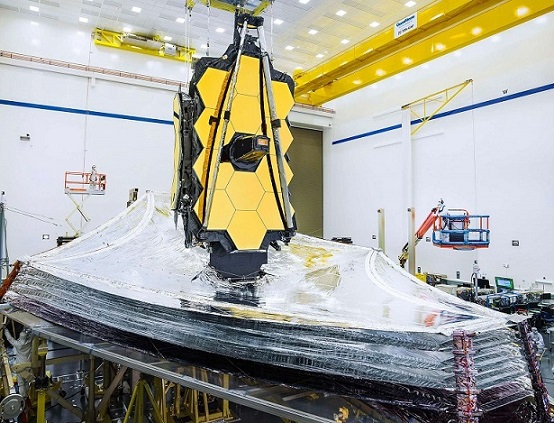 Photo en salle blanche du JWST partiellement déployé au-dessus de son bouclier thermique : celui-ci est en position presque horizontale et ressemble à un cerf-volant géant, recouvert d'une couche aux reflets argentés. 