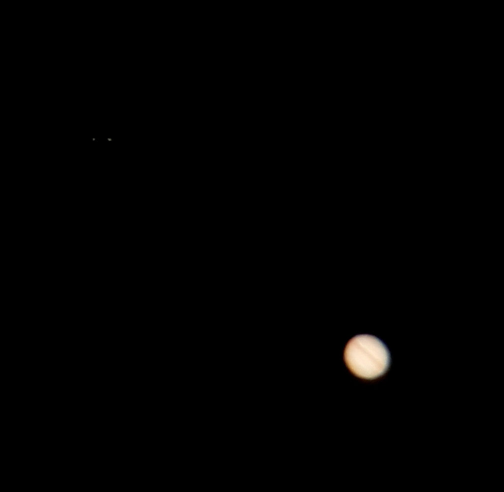 Photo de la planète Jupiter dont on voit le globe en bas et à droite de l'image. Deux petits points en haut et à gauche sont deux satellites de la planète. Sur le globe de Jupiter, on voit des bandes orangées de nuages. Le reste du globe est beige.