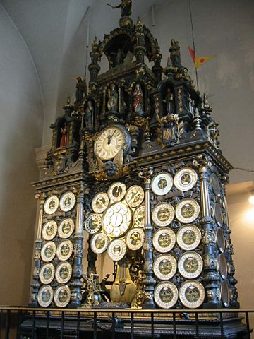 Photo de l'horloge de Besançon : elle comporte deux fois deux colonnes de cinq cadrans sur les côtés, et au centre huit cadrans disposés de façon circulaire. L'heure est en haut, et l'horloge est surplombée d'une structure scupltée qui pointe vers le ciel, comme un chapiteau. 