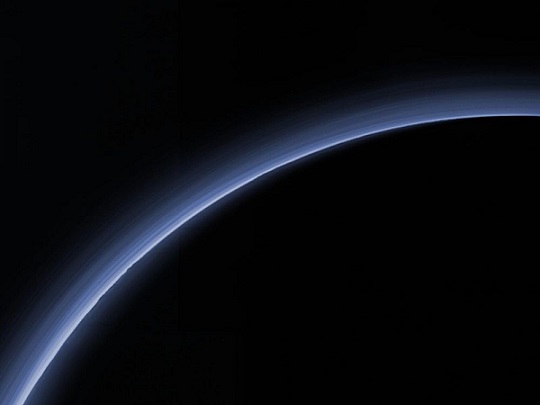 On voit en noir sur fond noir la courbe de la planète naine Pluton, soulignée par sa fine atmosphère, une couche bleutée. 