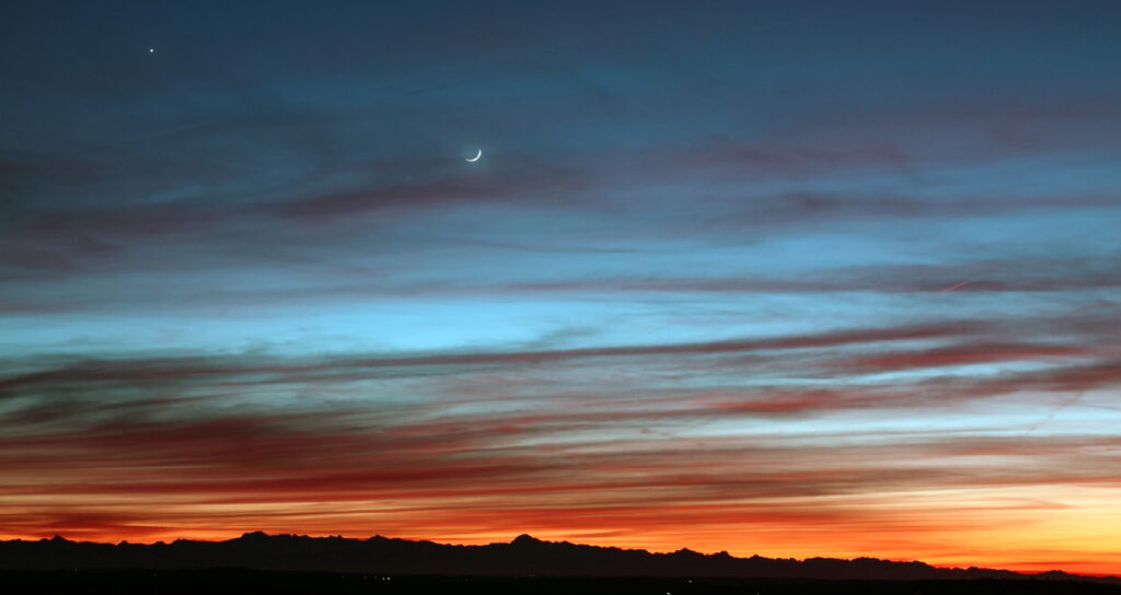 Photo montrant un crépuscule coloré du bleu au rouge, avec à l'horizon une chaine de montagne. Dans le ciel voilé de cirrus, on voit le fin croissant de lune et la planète Vénus sous la forme d'un point brillant en haut et à gauche de l'image.