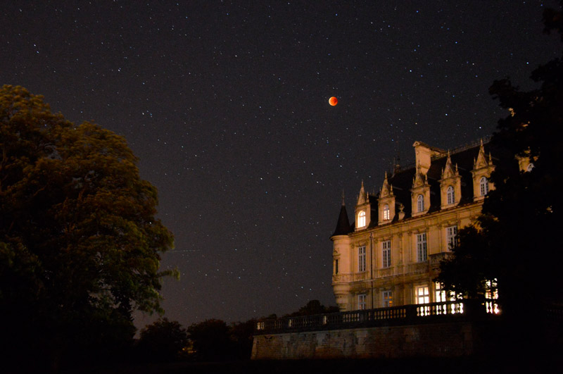 Photo montrant une éclipse de lune dans sa phase totale. La Lune est orange, le ciel est étoilée et en premier plan on a un château de type renaissance éclairé et des arbres en ombre chinoise.