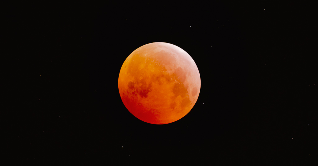 Photo montrant la lune complêtement orange, avec un c^té plus sombre et un côté plus clair. La couleur est due à l'éclipse de lune. En arrière-plan, le ciel noir et quelques étoiles.