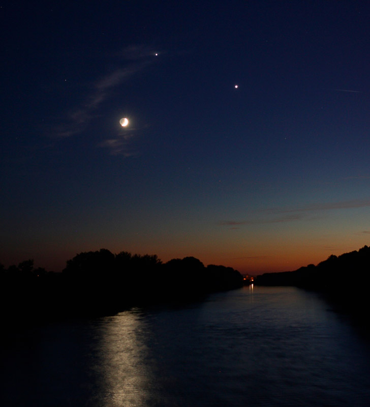 Image d'un crépuscule au dessus d'une rivière, le ciel est bleu en haut, orangée près de l'horizon. La rivière est bordée d'arbres. Un croissant de lune avec lumière cendrée est visible et au dessus il y a deux points lumineux qui sont les planètes Vénus et Jupiter.