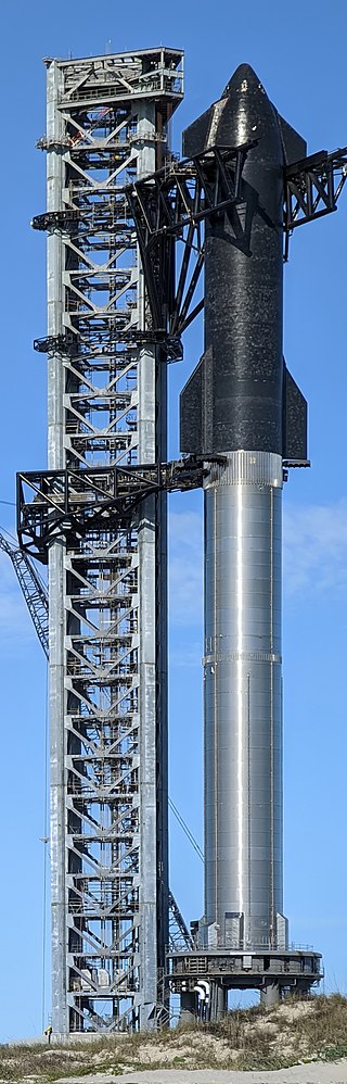 Sur fond de ciel bleu, une fusée Starship de couleur gris foncé sur un pas de tir, en cours de développement. 