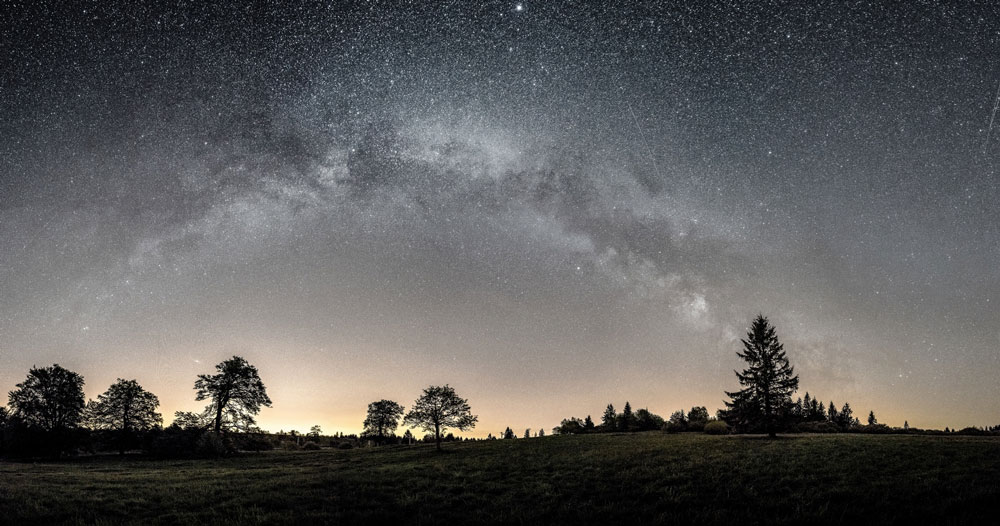 Photo de ciel étoilé montrant l'arche de la Voie lactée. En premier-plan, un espace plat et dégagé agrémenté d'arbres.