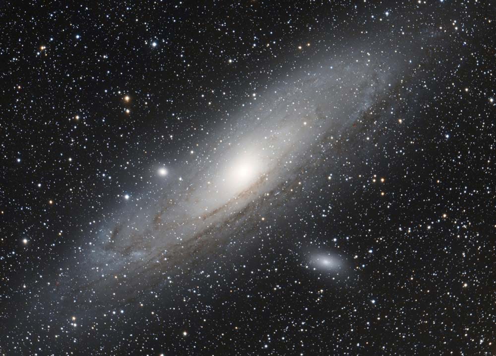 La galaxie M31 ressemble à un disque vu de 3/4. Elle est blanche et comporte des bandes de poussières sombres. EN bas à droite, on voit M110 et en haut à gauche M82 qui sont deux petites galaxies satellites.