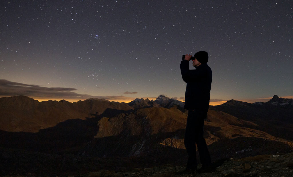 Image montrant un observateur debout tenant une paire de jumelles. Il regarde le ciel étoilé au dessus des montagnes.