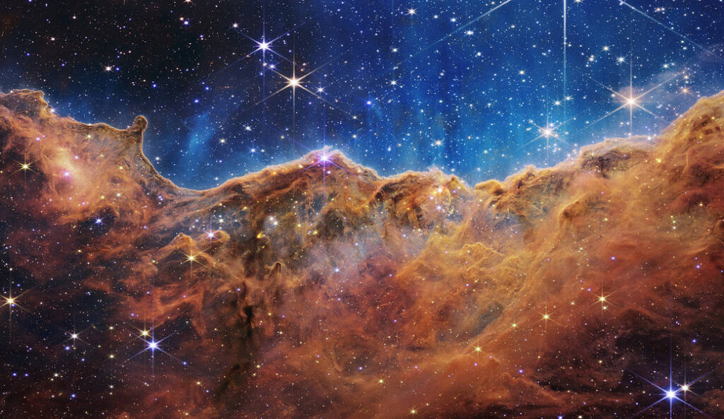 Des centaines de milliers d'étoiles dans la nébuleuse de la Carène. Grand nuage de gaz et de poussière marron-orange sur les deux tiers inférieurs de l'écran, et le tiers supérieur est bleuté.