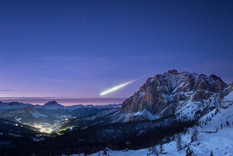 En bas de l'image les montagnes enneigées et une vallée, le ciel est violet légèrement rosé, et on voit une grosse étoile filante qui se dirige de la droite vers la gauche de l'image.