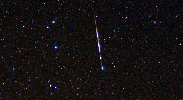 Sur fond de ciel étoilé, une étoile filante, longue ligne brillante, du haut vers le bas de l'image. 