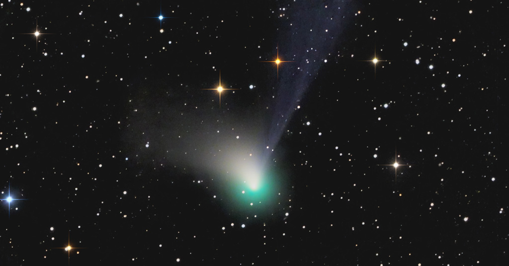 Comète C/2022 E3 (ZTF) avec la queue de gaz bleue qui part à la verticale, la queue de poussières blanchevers la gauche et le halo bleu-vert. Le tout sur un fond étoilé