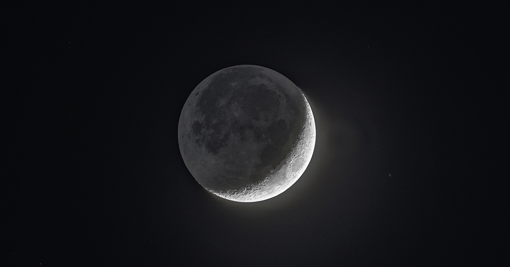 Photo de la Lune en gros plan, avec un croissant en bas à droite et la lumière sendrée sur le reste du globe où on distingue les mers et océans lunaires comme des zones plus sombres.