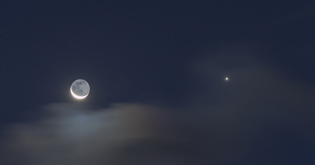 Photo de la Lune en fin croissant avec la lumière cendrée à gauche de l'image, et à droite l'étincelante planète Vénus. Un fin voile nuageux est visible en bas à gauche, sur un fond de ciel bleu nuit.