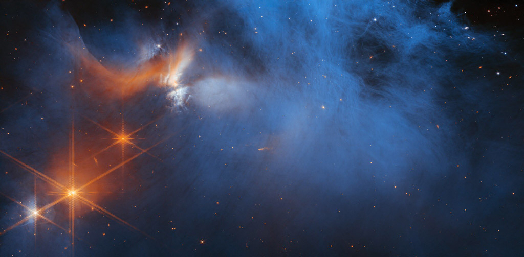 Image de la région centrale du nuage moléculaire sombre Chamaeleon I, qui montre principalement des traînées gazeuses bleues sur un fond sombre. Le côté supérieur gauche comporte en outre des nuages orange et blancs. Juste en dessous d'eux se trouvent quatre points de lumière brillants. Trois sont orange et un est un mélange de blanc et d'orange. Chacun de ces points a les pics de diffraction oranges à 8 points générés par l'optique du télescope James Webb. Dispersées sur toute l'image se trouvent des étoiles ou des galaxies lointaines dans des tons de rouge, d'orange et de bleu, vues comme de minuscules gouttes.