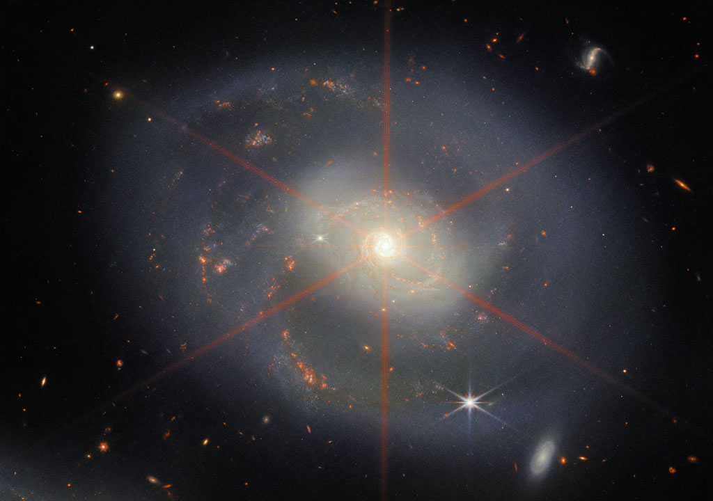Cette image montre une galaxie spirale dominée par une région centrale brillante. La galaxie a des teintes bleu-violet avec des régions orange-rouge remplies d'étoiles. Un grand pic de diffraction est également visible, qui apparaît comme un motif en étoile au-dessus de la région centrale de la galaxie. Beaucoup d'étoiles et de galaxies remplissent la scène d'arrière-plan.