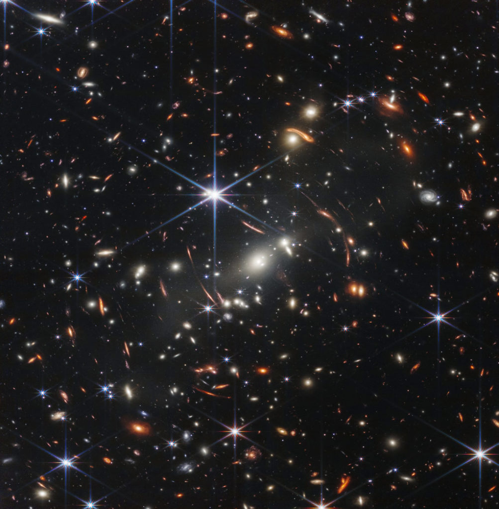Le fond de l'espace est noir. Des milliers de galaxies apparaissent partout dans l'image. Leurs formes et leurs couleurs varient. Certaines sont de différentes nuances d'orange, d'autres sont blanches. La plupart des étoiles apparaissent bleues et sont parfois aussi grandes que des galaxies plus éloignées qui apparaissent à côté d'elles. Une étoile très brillante est juste au-dessus et à gauche du centre. Elle a huit longs pics de diffraction bleu vif. Entre 4 heures et 6 heures dans ses pointes se trouvent plusieurs galaxies très brillantes. Un groupe de trois est au milieu et deux sont plus proches de 4 heures. Ces galaxies font partie de l'amas de galaxies SMACS 0723, et elles déforment l'apparence des galaxies vues autour d'elles. De longs arcs orange apparaissent à gauche et à droite vers le centre.
